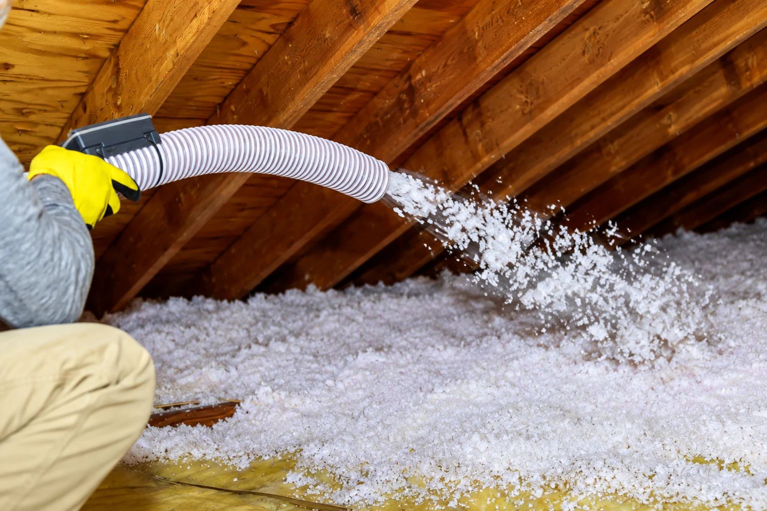 A worker directs a hose that blows loose white insulation across an attic floor.