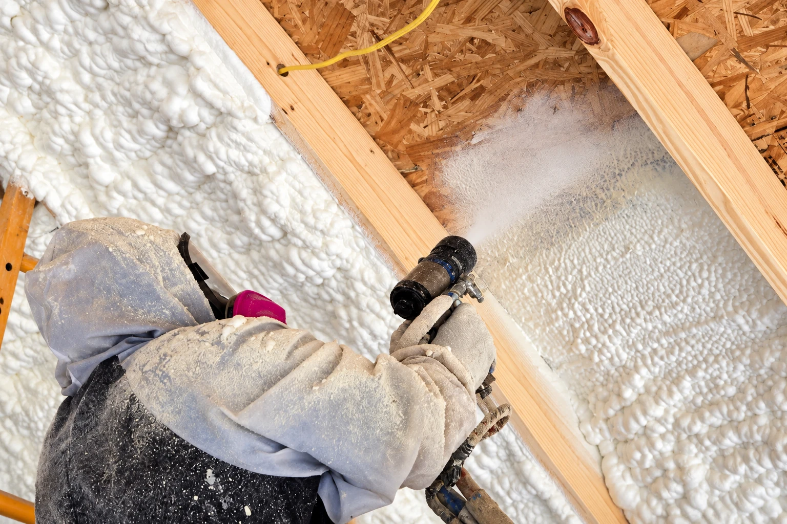 A worker in protective clothing sprays closed-cell foam insulation into a wooden wall cavity.