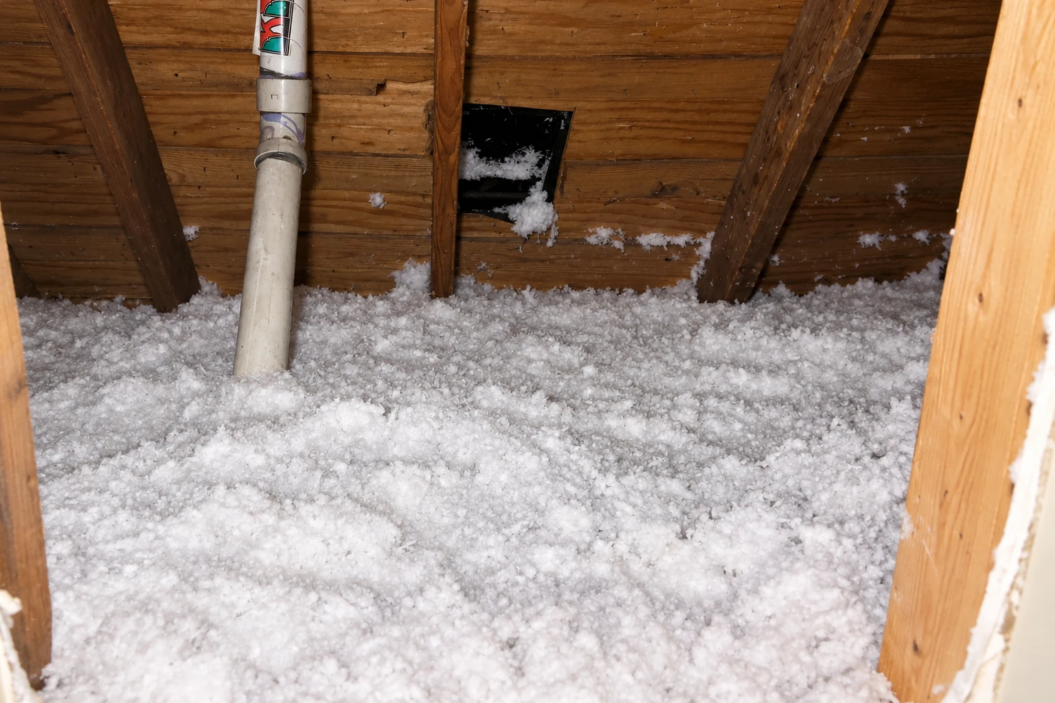 Attic floor is covered with loose white blown-in insulation around a pipe and wooden rafters.