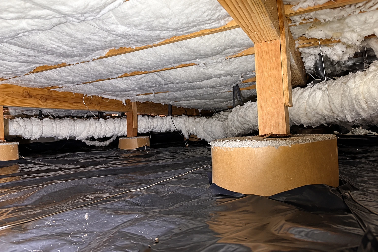 Crawl space has white insulation beneath floor joists and across ducts, above a black plastic vapor barrier.