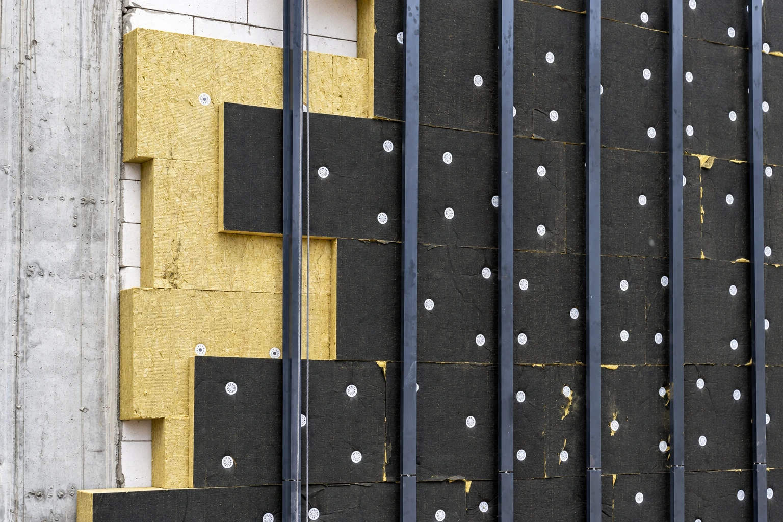 Black insulation boards are fastened over yellow wall insulation beside a concrete wall.